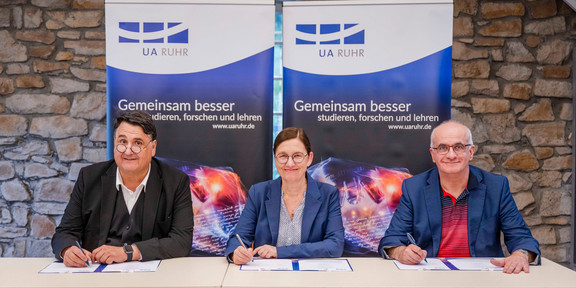 Foto: Die drei Rektor*innen der Universitäten der UA Ruhr sitzen an einem Tisch und unterzeichnen Dokumente.
