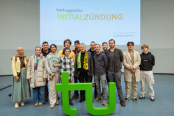 Prof. Dr. Klaus von Klitzing steht mit einer Gruppe von Teilnehmenden vor dem grünen TU Logo.