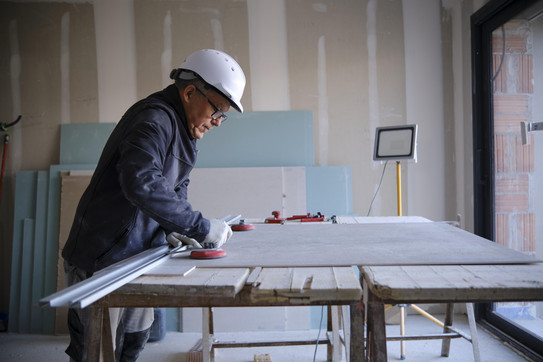 Ein älterer Mann, der einen Schutzhelm und Arbeitsmontur trägt, steht an einem Tisch und arbeitet mit Werkzeug an einer dünnen Holzplatte. 