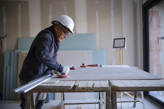 Ein älterer Mann, der einen Schutzhelm und Arbeitsmontur trägt, steht an einem Tisch und arbeitet mit Werkzeug an einer dünnen Holzplatte.