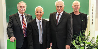 Four gentlemen in dark suits stand behind a large TU logo and smile at the camera