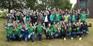 Gruppenfoto auf einer Wiese mit Bäumen und einem Gebäude im Hintergrund. 