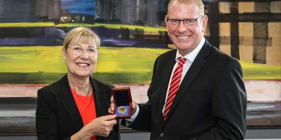 Rektorin Ursula Gather erhält eine Goldmedaille von Herrn Schaufelberger. Beide halten die Schatulle mit der Medaille fest und lächeln in die Kamera.