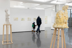 Ausstellung Goldenes Wunder Ein Mann mit Kind an der Hand geht durch eine Kunstausstellung. Rechts und links stehen Skulpturen auf Podesten aus, an den Wänden hängen Bilder.