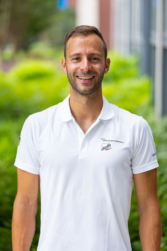 Portrait photo of Christoph Edeler: A photo of Christoph Edeler in a white polo shirt from the FISU Games. He stands in front of a blurred background.