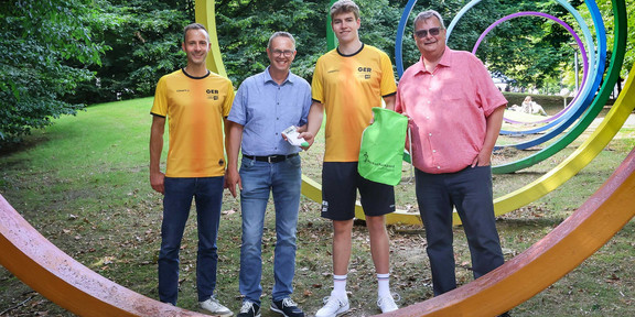 Fünf Männer mit verschiedenfarbigen Shirts und Hemden stehen hinter dem ersten Spektralring auf dem Campus der TU Dortmund. Der jüngste in der Runde bekommt kleine Präsente überreicht. 