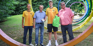 Fünf Männer mit verschiedenfarbigen Shirts und Hemden stehen hinter dem ersten Spektralring auf dem Campus der TU Dortmund. Der jüngste in der Runde bekommt kleine Präsente überreicht.