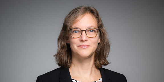 Porträtfoto von Prof. Nele McElvany: Prof. McElvany trägt einen dunklen Blazer und eine Brille. Sie steht vor einem grauen Hintergrund und lächelt in die Kamera.