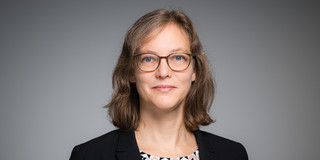Porträtfoto von Prof. Nele McElvany: Prof. McElvany trägt einen dunklen Blazer und eine Brille. Sie steht vor einem grauen Hintergrund und lächelt in die Kamera.