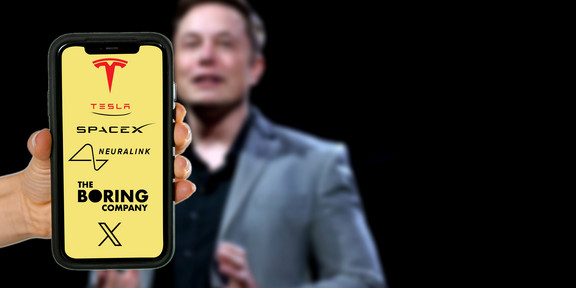 Multi-CEO Elon Musk Im Hintergrund ein leicht verschwommenes Foto von Elon Musk auf einer Bühne. Im Vordergrund hält eine Hand ein Smartphone ins Bild, auf dem Display sind die Logos von Tesla, SpaceX, Neuralink, The Boring Company und X auf gelbem Hintergrund zu erkennen.