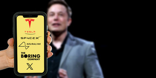 Multi-CEO Elon Musk Im Hintergrund ein leicht verschwommenes Foto von Elon Musk auf einer Bühne. Im Vordergrund hält eine Hand ein Smartphone ins Bild, auf dem Display sind die Logos von Tesla, SpaceX, Neuralink, The Boring Company und X auf gelbem Hintergrund zu erkennen.