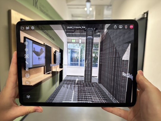 iBuild AR Zwei Hände halten einen Tablet-PC auf dem eine Augmented-Reality-App die unterliegenden Tragkonstruktionen des Raums zeigt, der hinter dem Tablet zu sehen ist.