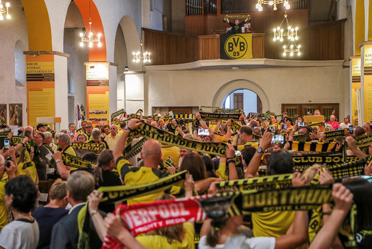 Auf dem Foto sieht man eine Kirche von innen, in der viele Menschen nebeneinander stehen und den BVB-Schal in die Höhe strecken. 