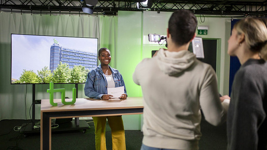 Moderatorin im Studio hinter Tisch mit „tu“-Logo, während ein Kamerateam sie filmt; auf dem Bildschirm hinter ihr ist ein Universitätsgebäude.