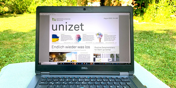 A laptop stands on a table that has been set up on a green area on the TU Dortmund campus. On the monitor of the laptop, a window with an issue of unizet is open.