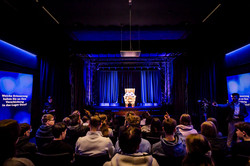Foto: Eine Gruppe von Menschen sitzt in einem dunklen Raum und betrachtet ein Hologramm auf einer beleuchteten Bühne mit einem Sessel im Zentrum.