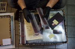 Zwei Hände mit schwarzen Handschuhen sortieren durch einen Stapel Fotonegative über einem Karton, der durcheinander mit Fotos, Negativen und weiteren Dokumenten gefüllt ist.