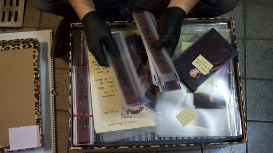 Zwei Hände mit schwarzen Handschuhen sortieren durch einen Stapel Fotonegative über einem Karton, der durcheinander mit Fotos, Negativen und weiteren Dokumenten gefüllt ist.