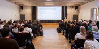 Das Bild zeigt einen Saal, an den Seiten Publikum und im Zentrum ein Gang, welcher nach Vorne zur Leinwand führt.
