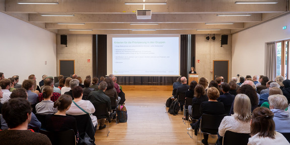 Das Bild zeigt einen Saal, an den Seiten Publikum und im Zentrum ein Gang, welcher nach Vorne zur Leinwand führt.