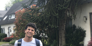 Ein Mann steht vor einem Baum und Häusern und lächelt in die Kamera