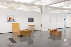 In einer Ausstellung stehen verschiedene Ausstellungsobjekte wie Bilder und Holzkästen vor einer weißen Wand.