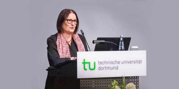 Dr. Irina Scherbakowa hinter einem Rednerpult mit dem Logo der TU Dortmund. Der Hintergrund ist grau.