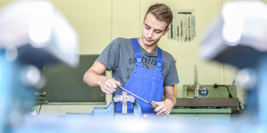 A young man in a blue overall working at a workbench.
