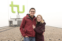 Ein Mann und eine Frau lächeln in die Kamera. Sie stehen auf dem Dach des Mathetowers vor dem TU-Logo.