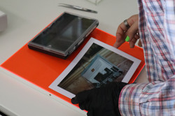 Eine Person in einem blau-rot karierten Hemd steht vor einem weißen Tisch und legt vorsichtig mit einem schwarzen Handschuh ein Foto und eine Fotobox auf eine neonorange Platte.