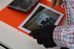 A person in a blue and red checked shirt stands in front of a white table and carefully places a photo and a photo box on a neon orange plate with a black glove.