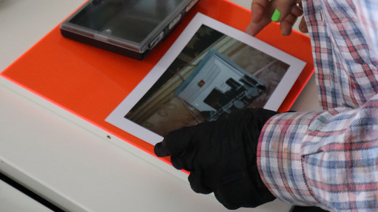 Eine Person in einem blau-rot karierten Hemd steht vor einem weißen Tisch und legt vorsichtig mit einem schwarzen Handschuh ein Foto und eine Fotobox auf eine neonorange Platte.