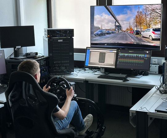 Foto: Eine Person sitzt an einem Fahrsimulator mit mehreren Monitoren und steuert virtuell ein Fahrzeug auf einer Straße.