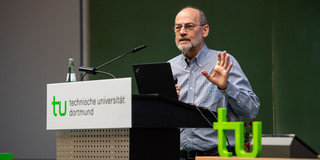 Das Bild zeigt eine männlich gelesene Person hinter einem Pult. Vorn am Pult hängt das TU-Logo-Schild. Das grüne TU-Logo als "Aufsteller" ist rechts unten im Bild zu sehen.