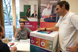 Infostand von viadee auf der Internationalen Karrieremesse Ein Mann sitzt an einem Infostand, während ihm ein weiterer Mann gegenüber sitzt. Hinter dem Tisch sind zwei Aufsteller der Firma viadee zu sehen. Im Vordergrund steht ein weiterer Mann, der ein T-Shirt mit dem Logo der Firma viadee trägt.