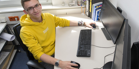 A young man in a yellow sweater sits at a desk with a computer and holds the mouse with his right hand
