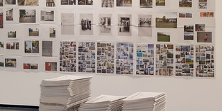 Fotos hängen in einer Ausstellung an der Wand, im Vordergrund liegt ein Stapel Zeitungen
