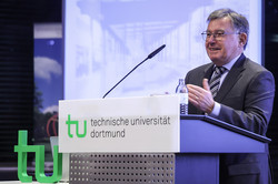 Foto: Eine Person spricht am Rednerpult der Technischen Universität Dortmund vor einer Leinwand mit einer Präsentation.
