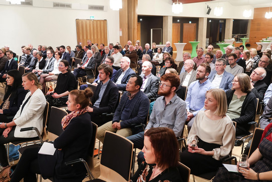 The audience of a seated event, all have their eyes straight ahead (directed to the left edge of the picture). In the back of the room, standing tables can be seen.