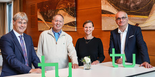 Prof. Quante, Prof. Barzilai, Frau Prof. Flatten und Rektor Prof. Bayer stehen gemeinsam an einem Tisch, auf dem das TU-Logo steht.