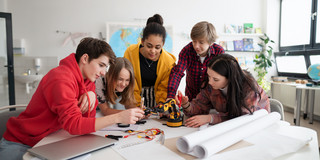 Eine Gruppe von Jugendlichen steht um einen Tisch herum. Alle beugen sich über einen kleinen Roboterarm. Ein Mädchen schraubt an dem Roboterarm. Im Vordergrund liegen Papierrollen und ein Laptop.