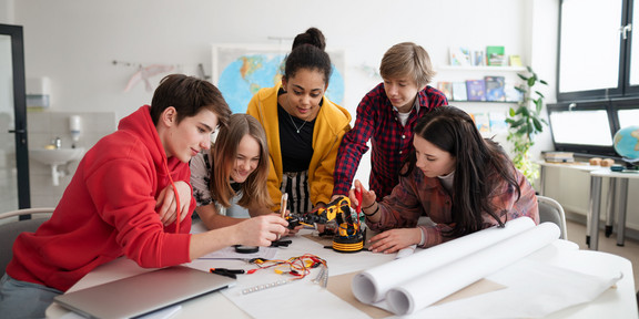 Eine Gruppe von Jugendlichen steht um einen Tisch herum. Alle beugen sich über einen kleinen Roboterarm. Ein Mädchen schraubt an dem Roboterarm. Im Vordergrund liegen Papierrollen und ein Laptop.