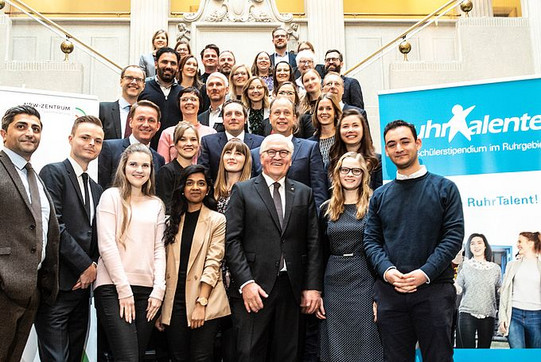 Eine Gruppe von Menschen, darunter auch der Bundespräsident Frank-Walter Steinmeier, befindet sich auf der Treppe eines festlichen Gebäudes. Rechts daneben steht ein blau-weißes Banner des RuhrTalente-Stipendienprogramms.