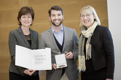 Eine Frau, ein Mann und eine Frau stehen neben einander. Die Frau links hält eine Urkunde in den Händen, der Mann hält eine Medaille in einem Kästchen in den Händen.