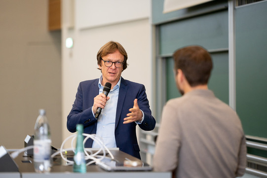 Auf dem Foto sieht man zwei Personen vorne im Hörsaal sprechen. Im Fokus ist ein Mann, der ein Mikrofron in der Hand hält und spricht.