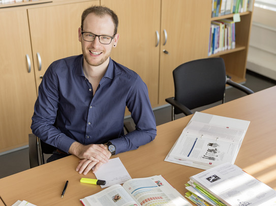 Foto von einem Mann und er sitzt an einem Tisch mit Büchern, Stiften und Zetteln vor ihm.