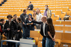 Mehrere Personen stehen in einem Hörsaal. Im Vordergrund schießt ein Mann mit einem Handy ein Selfie mit einer jungen Frau.