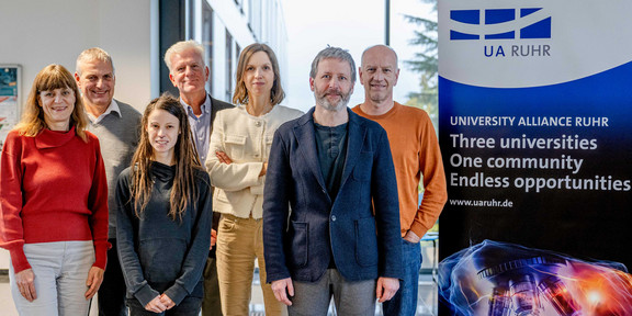 Foto: Ein Gruppenfoto vom UA Ruhr Rat für Ethik und Verantwortung neben einem Aufsteller der UA Ruhr. Auf dem Aufsteller steht: "University Alliance Ruhr. Three universities, one community, endless opportunities""