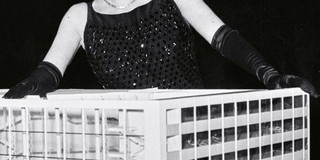 Eine Frau in einem schicken Kleid posiert hinter einem architektonischen Modell eines Gebäudes.