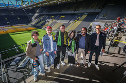 Auf dem Foto sieht man sechs Personen auf der Tribüne stehen und für ein Foto posieren. Alle lächeln in die Kamera.